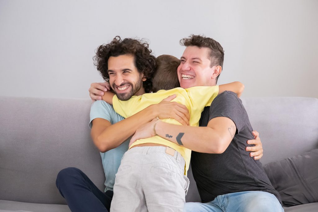 Pareja de hombres sonriendo y abrazando a su hijo en un sofá, reflejando la subrogación como una opción ideal en diversas situaciones.