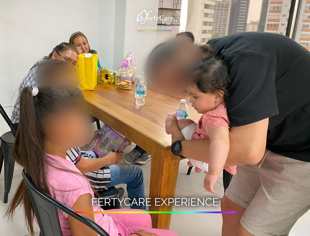Un padre acerca a su bebé a una niña mientras comparten un momento familiar alrededor de una mesa, representando la experiencia de subrogación en FertyCare.