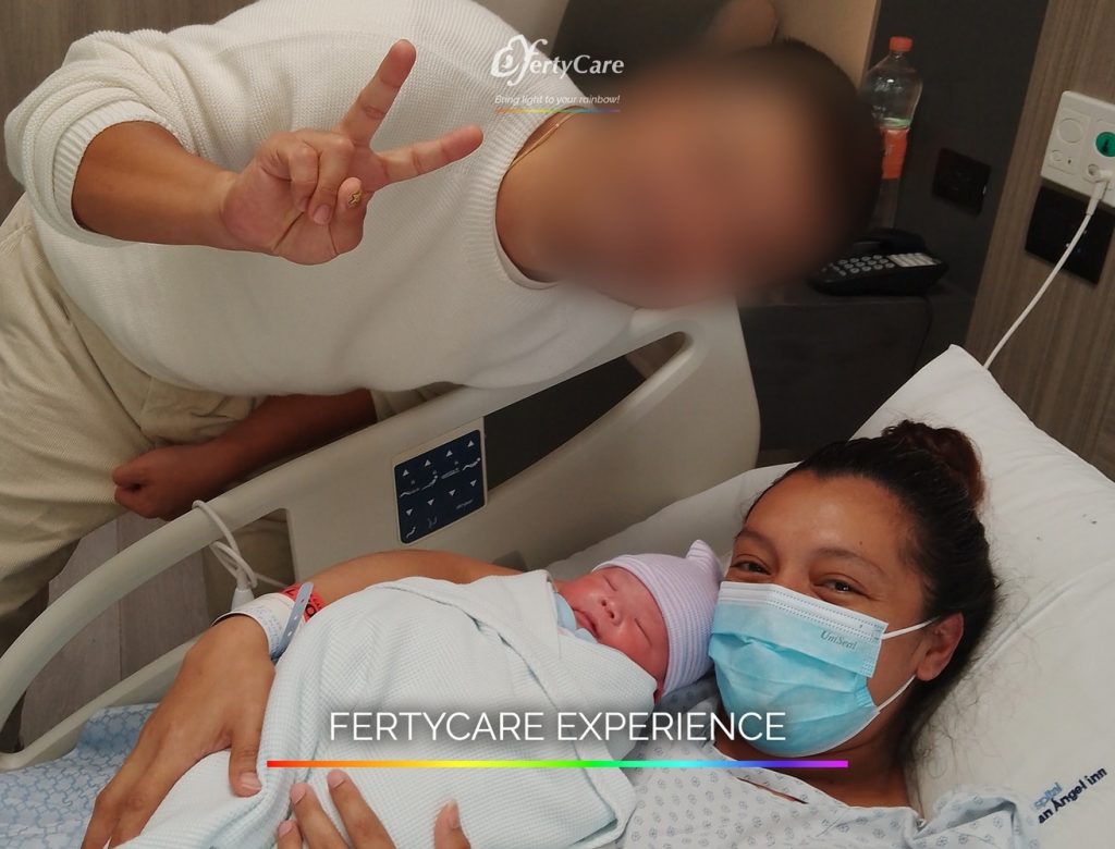 Madre sosteniendo a su bebé recién nacido mientras su acompañante hace la señal de paz, representando la experiencia de subrogación en FertyCare.