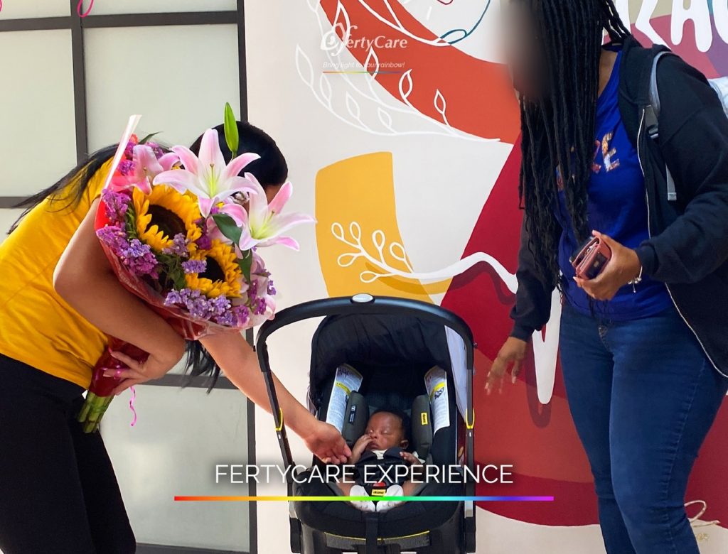 Dos mujeres, una sosteniendo un ramo de flores, saludan a un bebé en un portabebés dentro de las oficinas de FertyCare, representando la experiencia de subrogación.